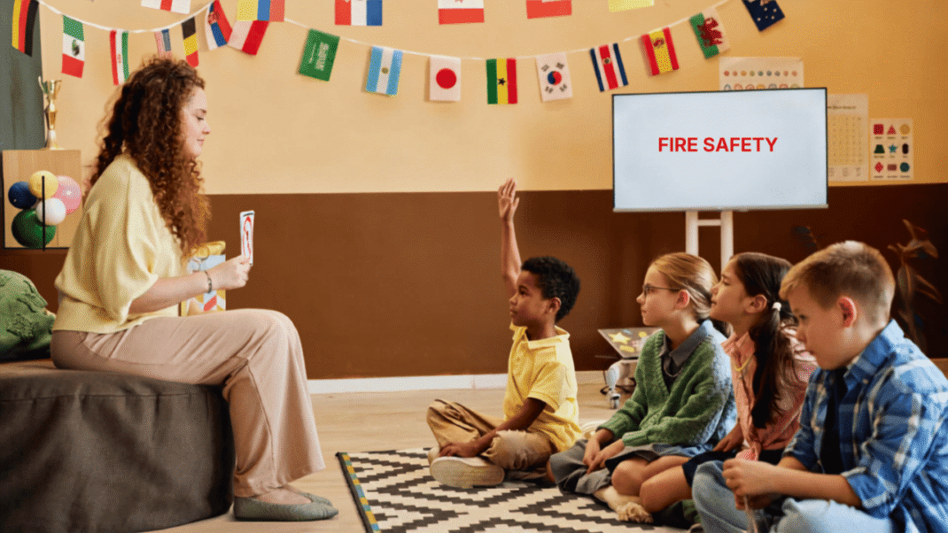 A teacher sits with four children in a classroom, one child raising a hand. A screen displays “FIRE SAFETY” and flags hang on the wall in the background.
